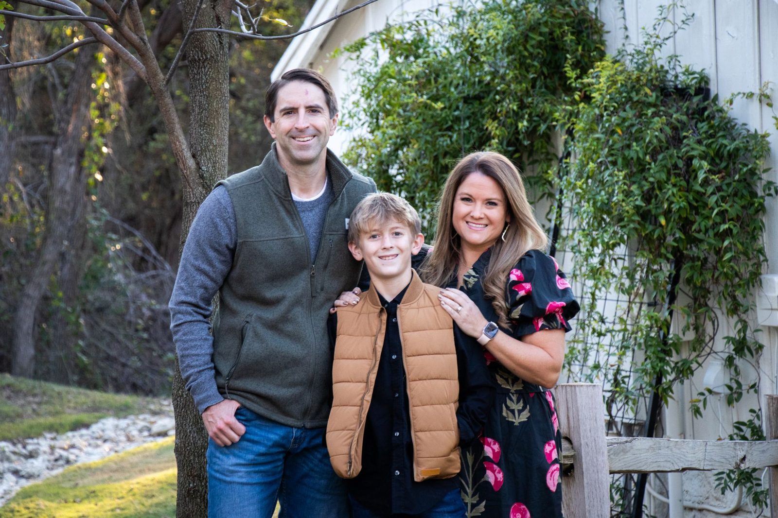 Family of three smiling by a white barn and a wooden fence